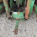Rusty green farm implement frame with hydraulic hoses and bolts on a gravel surface, showing wear and rust.