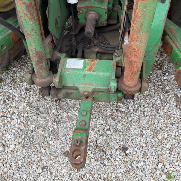 Rusty green farm implement frame with hydraulic hoses and bolts on a gravel surface, showing wear and rust.