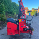 Red hydraulic construction machine on a street with a hedge on the left and brick houses in the background.