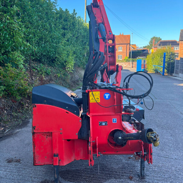 Red hydraulic construction machine on a street with a hedge on the left and brick houses in the background.