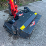 Red hydraulic arm of a KUHN mower attachment resting on a dark blue deck on an asphalt surface with yellow warning labels nearby, outdoors.