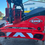 Close-up of red KUHN agricultural machine with hydraulic hoses and metal frame, 'POLY-LONGER' branding visible in foreground.