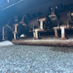 Underbelly of a freight train car showing rusted metal brackets, pins, and chains along the chassis near a rusted pipe, with a concrete ground below.