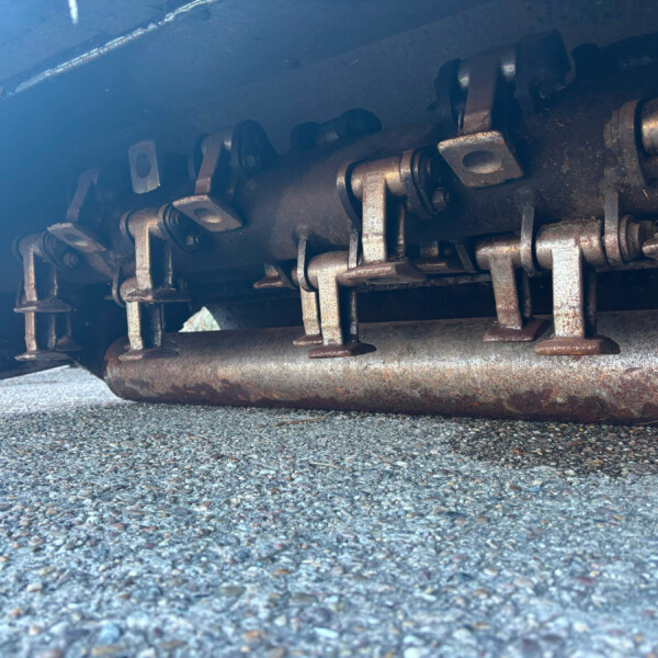 Underbelly of a freight train car showing rusted metal brackets, pins, and chains along the chassis near a rusted pipe, with a concrete ground below.