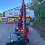 Red hydraulic agricultural machine with extended arms parked in front of a New Holland Agriculture storefront.