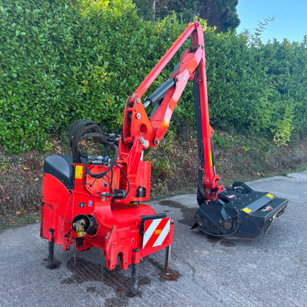 Red compact excavator with hydraulic arm raised, parked beside a tall green hedge on a sunny day near a sidewalk.