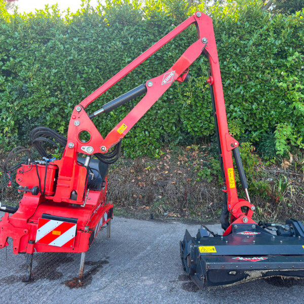 Red hydraulic construction crane attachment mounted on a small vehicle, set against a dense green hedge.