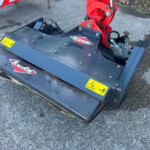 Close-up of a Kuhn agricultural mower attachment connected to a red hydraulic arm, sitting on a gravel surface with yellow safety stickers visible.