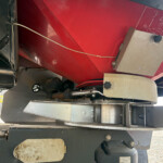 Underbelly of heavy industrial equipment: red chassis with metal panels, exposed wiring, and a gray control/housing unit nearby.