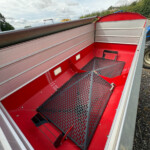 Red utility trailer interior with perforated metal ramp and seating steps, viewed from the open side, outdoors on gravel.