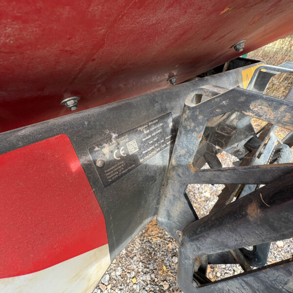 Close-up of a red boat hull attached to a black metal trailer, showing bolts along the hull and a dusty, rocky ground beneath.