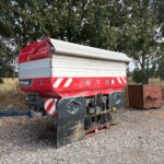 Red agricultural trailer with a white roll-up cover parked on gravel, behind a wire fence with trees in the background.