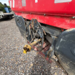 Red boat on a trailer with tangled ropes and a yellow hitch resting on a gravel lot, with blue containers and tires in the background.