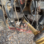Close-up of a muddy trailer hitch area with tangled hoses, cables, and a metal chain under a dusty vehicle.