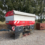 Red agricultural trailer with a white rolled tarp cover, parked on gravel near a metal fence and trees in the background.