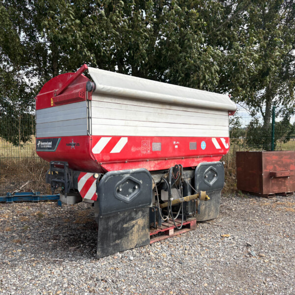 Red agricultural trailer with a white rolled tarp cover, parked on gravel near a metal fence and trees in the background.