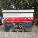 Blue tractor attached to a large red-and-silver trailer on a gravel lot with trees in the background.