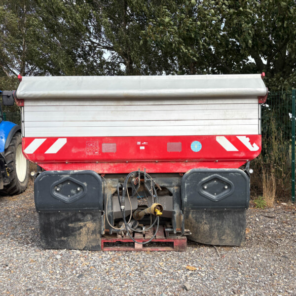 Blue tractor attached to a large red-and-silver trailer on a gravel lot with trees in the background.