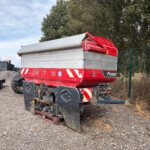 Red and silver fertilizer spreader trailer in a gravel yard, with a green fence and trees in the background.
