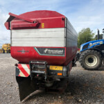Rear view of a red Kverneland TLX Geospread spreading trailer hitched to a blue New Holland tractor in a gravel yard, with logos and hazard stripes visible.