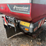 Rear view of a red agricultural trailer with Kverneland TLX GEOSPREAD branding and hazard stickers, parked on a gravel lot. It shows a metal hitch, steps, and a wooden pallet beneath the trailer.
