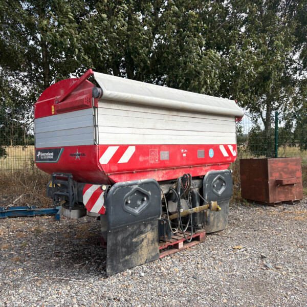 Red agricultural trailer with a white roll-up cover parked on gravel, behind a wire fence with trees in the background.