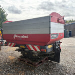 Red Kverneland agricultural machine mounted on a trailer, parked in a gravel lot beside a blue-roofed metal building under a cloudy sky