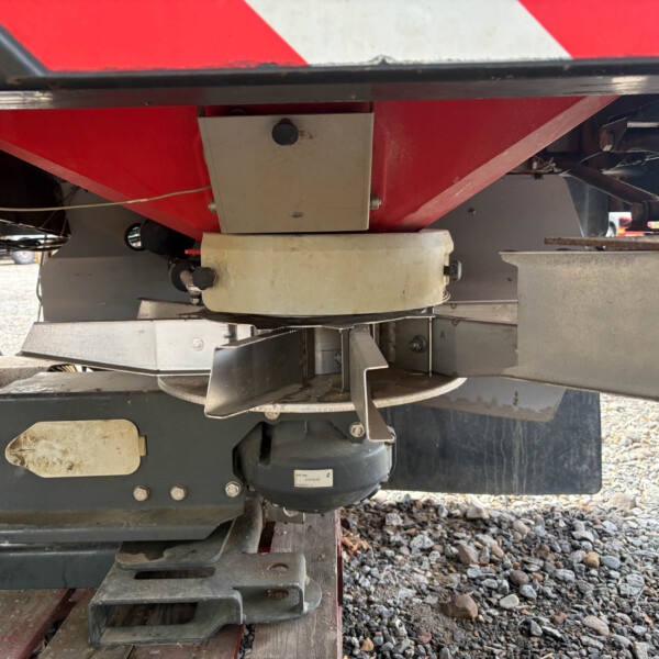 Underside of a red-painted trailer or machine with metal brackets, a gray rectangular cover, and a black motor, on a gravel surface.