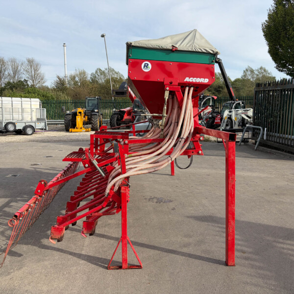 Red agricultural spreader with beige hoses trailing from the machine in an outdoor equipment yard. Nearby trailers and farm equipment are visible behind a fence.