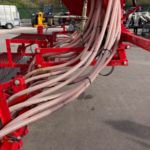 Red hydraulic agricultural equipment with many coiled hoses and a metal frame, parked in an outdoor yard with trailers in the background with a fence.