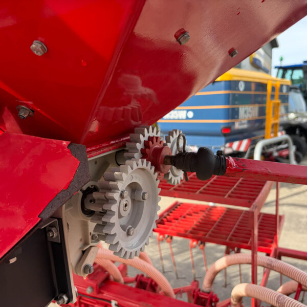 Close-up of red industrial machinery with gears, bolts and hydraulic hoses; a blue vehicle is blurred in the background.