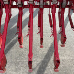 Row of red metal farm implement shanks with springs, resting on a concrete surface.