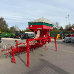Red agricultural seeding machine with green top covered by a beige canopy in a yard, surrounded by tractors and equipment.