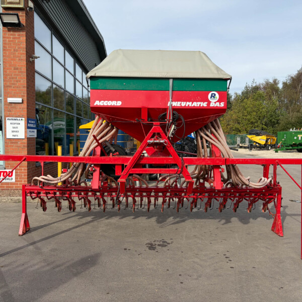 Red and green pneumatic seed drill with hoses mounted on a red frame outside a dealership building labeled ACCORD PNEUMATIC DAS, facing front-right.