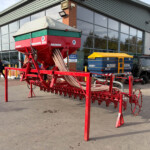 Red farm seeding machine on a trailer displayed outside a modern agricultural showroom with people nearby.