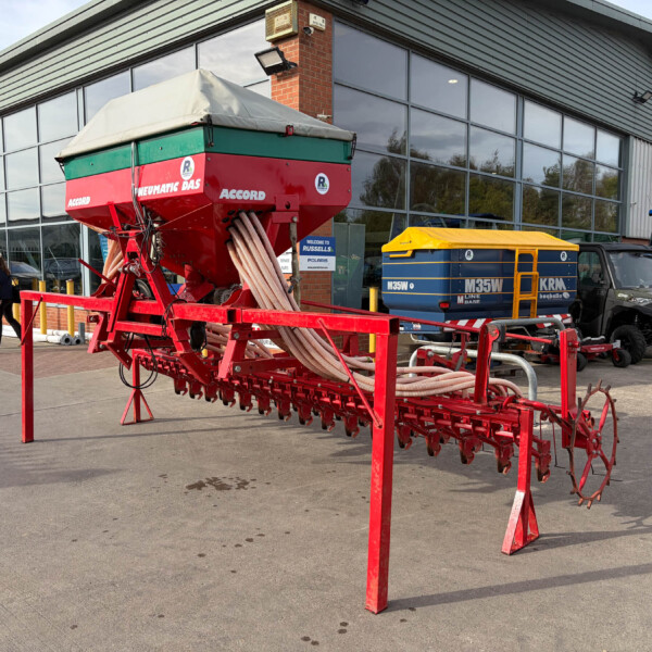 Red farm seeding machine on a trailer displayed outside a modern agricultural showroom with people nearby.