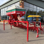 Red agricultural seeding machine on a trailer parked outside a glass-fronted industrial building; two pedestrians walk nearby.
