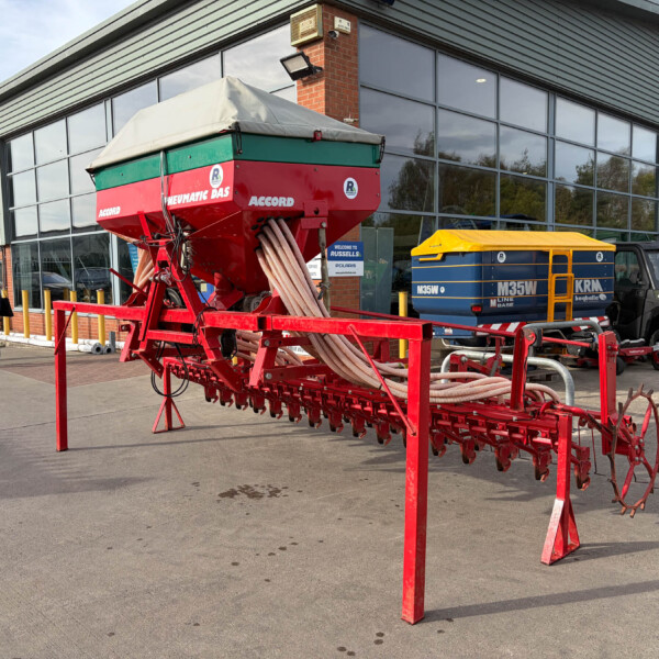 Red agricultural seeding machine on a trailer parked outside a glass-fronted industrial building; two pedestrians walk nearby.