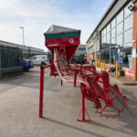 Red agricultural seed planter outside a showroom with large glass windows and brick facade; hoses hang from the equipment.