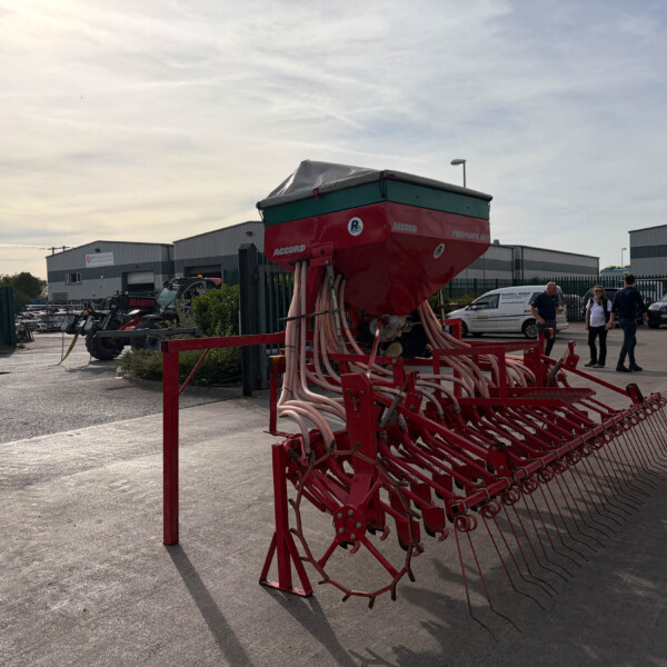 Large red agricultural seed drill with hoses in a parking lot near an industrial complex; a few people and cars in the background.