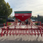 Front view of a red pneumatic seed drill with teal top, many pink hoses hanging from its frame, parked behind a fence in an outdoor lot.