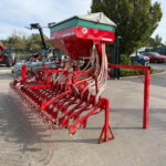 Red agricultural planter machine with a large red hopper and beige cover, hoses trailing along a red frame on a paved lot with tractors in the background.