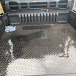 Diamond-plate truck bed with ribbed side walls and a small blue cloth in the left corner. It’s the cargo area of a rugged utility vehicle, viewed from above.