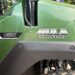 Close-up of a green tractor/utility vehicle side panel with a black hydraulic strut and a coiled cable, featuring the 'MULE PRO-DX' decal and black step area nearby.