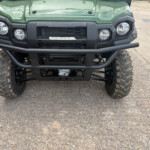 Front view of a green Kawasaki off-road vehicle with a rugged black bumper, round headlights, and large knobby tires; a taped paper plate reads 00033 on the grille.