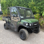 Green and black Kawasaki Mule utility vehicle parked on a paved lot with a tall hedge in the background, front-facing view showing its cab and tires.