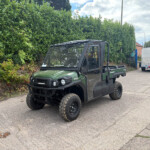 Front-side view of a green Kawasaki off-road utility vehicle parked along a hedge-lined street with a silver trailer in the background