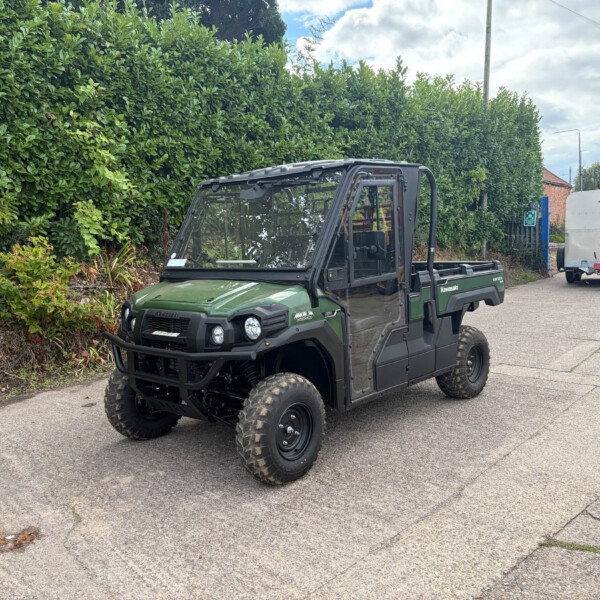 Front-side view of a green Kawasaki off-road utility vehicle parked along a hedge-lined street with a silver trailer in the background