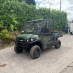 Green Kawasaki utility terrain vehicle parked on a paved street beside a tall hedge with a metal trailer in the background.