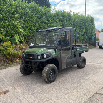 Green Kawasaki utility terrain vehicle parked on a paved street beside a tall hedge with a metal trailer in the background.